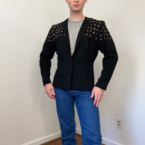 May include: A black blazer with gold and pink embellishments on the shoulders. The blazer has a fitted silhouette and a single button closure.