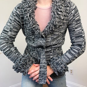 Könnte beinhalten: Schwarz-weiß gemusterte Strickjacke mit strukturiertem, gefranstem Kragen und Manschetten. Der Pullover hat einen passenden Gürtel und einen Knopfverschluss. Die Ärmel sind lang und das Gesamtdesign ist eine Mischung aus Streifen und strukturiertem Strick.