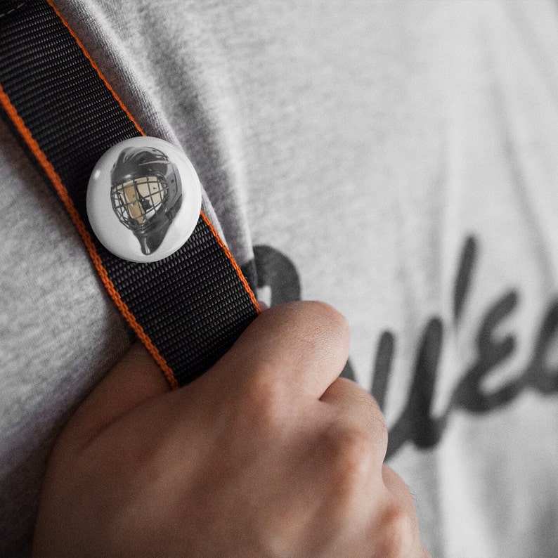 May include: A white button with a black and white image of a hockey helmet. The button is attached to a black and orange strap.
