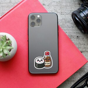 May include: A gray iPhone with a sticker of a cartoon sushi roll and a bottle of soy sauce. The sticker says "Soy Sauce".