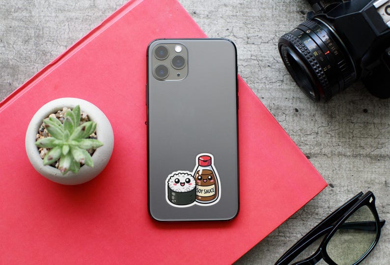 May include: A gray iPhone with a sticker of a cartoon sushi roll and a bottle of soy sauce. The sticker says "Soy Sauce".