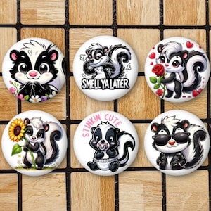 May include: Six round white buttons with black and white cartoon skunk illustrations. The buttons feature different poses and sayings, including "Smell Ya Later" and "Stinkin' Cute".