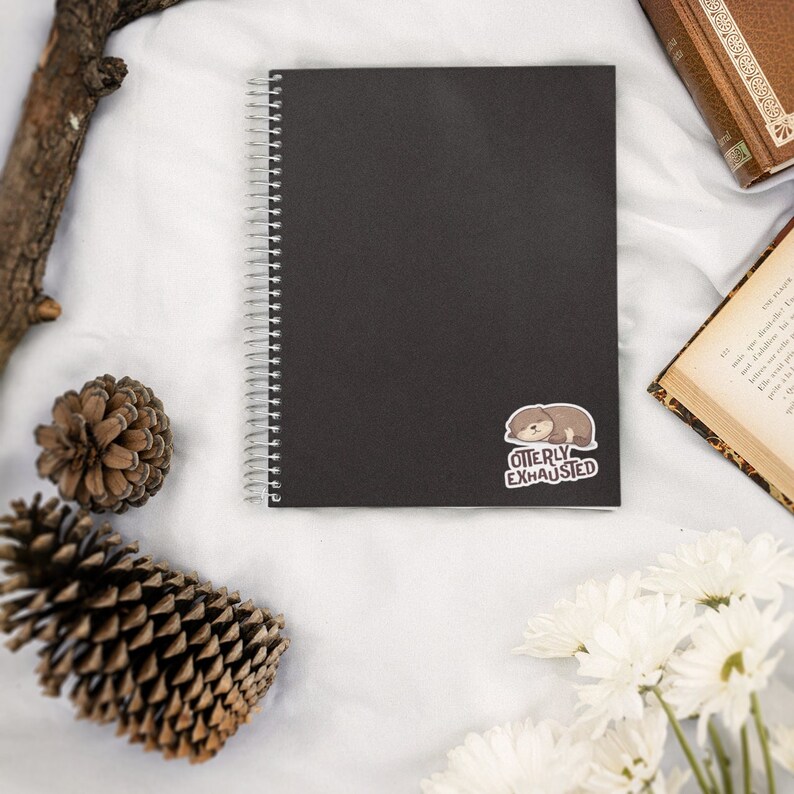 Op de afbeelding: Een zwarte spiraalgebonden notitieboek met een sticker van een slapende cartoon-otter en de tekst "Otterly Exhausted". Het notitieboek ligt op een wit oppervlak met dennenappels en witte bloemen.