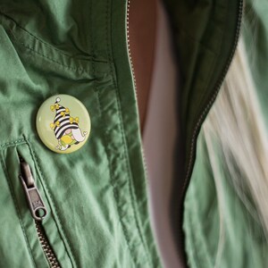 K&ouml;nnte beinhalten: Eine gr&uuml;ne Jacke mit Rei&szlig;verschluss und Knopf. Ein runder Knopf mit einem Cartoon-Zwerg, der einen schwarz-wei&szlig; gestreiften Hut mit gelben Zitronen tr&auml;gt, ist an der Jacke befestigt.