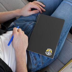 May include: A black spiral notebook with a yellow and black sticker that says "Trust me I'm a welder". The sticker features a cartoon image of a welder wearing a welding mask and holding a welding torch.