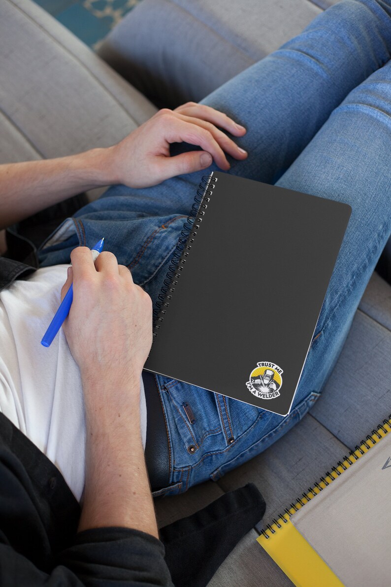 May include: A black spiral notebook with a yellow and black sticker that says "Trust me I'm a welder". The sticker features a cartoon image of a welder wearing a welding mask and holding a welding torch.