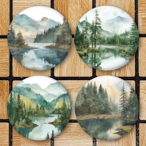 May include: Four round decorative coasters with watercolor landscape paintings. Each coaster features a different mountain and lake scene with trees in shades of green, blue, and brown. The coasters are arranged on a wooden surface.