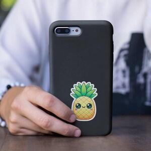 Pu&ograve; includere: Una custodia per iPhone nera con un adesivo di ananas a cartone animato. L'ananas &egrave; giallo con foglie verdi e ha un viso sorridente.