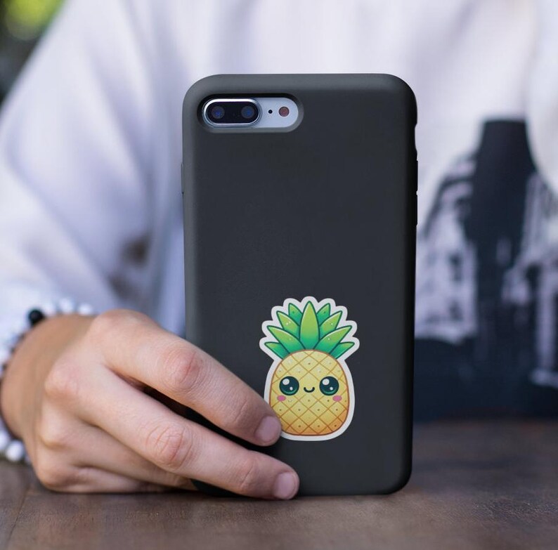 Pu&ograve; includere: Una custodia per iPhone nera con un adesivo di ananas a cartone animato. L'ananas &egrave; giallo con foglie verdi e ha un viso sorridente.