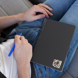 May include: A black spiral bound notebook with a sticker of a German Shepherd dog playing fetch with a tennis ball. The notebook is lying on a person's lap.