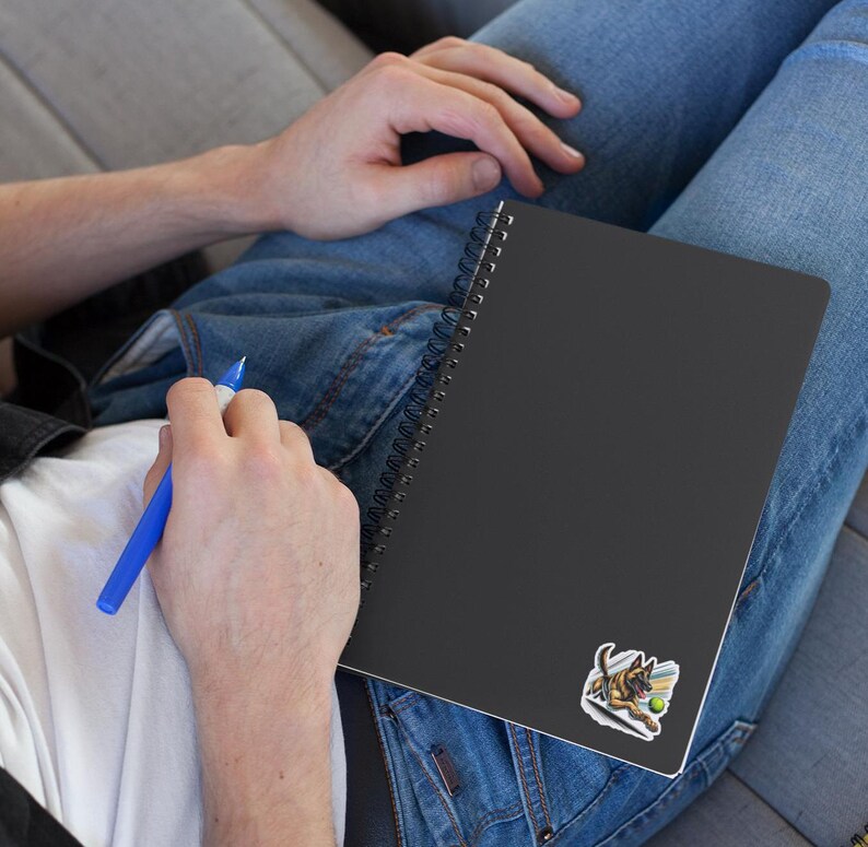 May include: A black spiral bound notebook with a sticker of a German Shepherd dog playing fetch with a tennis ball. The notebook is lying on a person's lap.