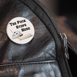 May include: A white button with a hockey mask, stick, and puck. The text on the button reads "The Puck Stops Here."