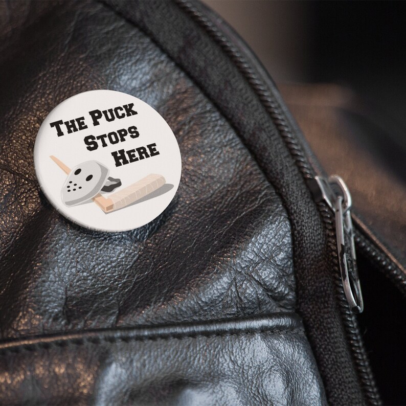 May include: A white button with a hockey mask, stick, and puck. The text on the button reads "The Puck Stops Here."