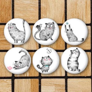 May include: Six white round buttons featuring various illustrations of grey cats. The cats are depicted in different poses, such as sitting, standing, and playing with a butterfly.