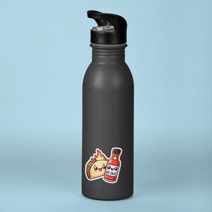 May include: A black reusable water bottle with a black lid and a sticker of a cartoon taco and hot sauce bottle. The taco and hot sauce bottle are both smiling and have flames coming out of them. The hot sauce bottle says "Hot Sauce" on it.