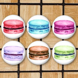 May include: A set of six circular buttons featuring watercolor illustrations of macarons in various colors including pink, blue, magenta, lavender, brown, and light green. Each button displays a single macaron with a visible filling layer.