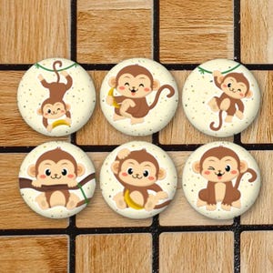 May include: A set of six circular buttons featuring cartoon monkey illustrations on a cream-coloured background with small brown speckles. Each button displays a different monkey pose, including monkeys hanging from vines and holding yellow bananas.