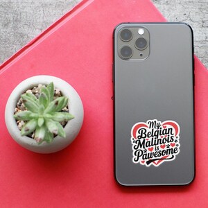 May include: A black iPhone with a white sticker that says "My Belgian Malinois is Pawsome" with a red heart and paw prints. The sticker is on the back of the phone.