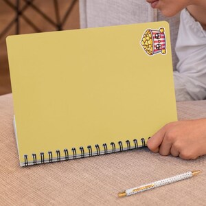 Pu&ograve; includere: Un quaderno giallo a spirale con un adesivo a forma di secchiello di popcorn di cartone animato sulla copertina anteriore. L'adesivo ha un viso carino ed &egrave; pieno di popcorn.