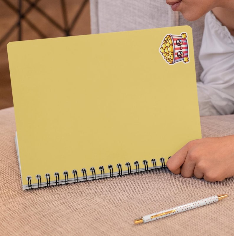 Pu&ograve; includere: Un quaderno giallo a spirale con un adesivo a forma di secchiello di popcorn di cartone animato sulla copertina anteriore. L'adesivo ha un viso carino ed &egrave; pieno di popcorn.
