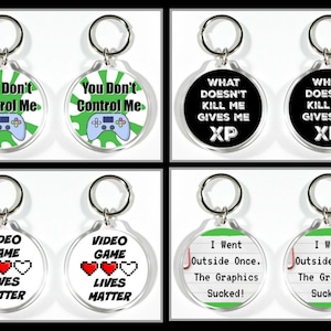 May include: Set of eight round keychains with different designs and sayings.  The keychains are made of clear acrylic with a white background.  The designs include a green and white video game controller with the text "You Don't Control Me", a black background with white text that says "What Doesn't Kill Me Gives Me XP", a white background with red hearts and the text "Video Game Lives Matter", and a green background with white text that says "I Went Outside Once. The Graphics Sucked!"