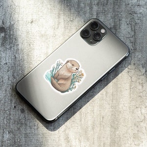 Op de afbeelding: Een sticker met een cartoon-otter met bruine vacht, omringd door gele bloemen op een blauwe achtergrond. De sticker zit op een zilveren telefoonhoesje.