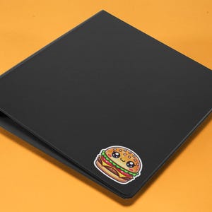 Pu&ograve; includere: Un raccoglitore nero con un adesivo di hamburger di cartone animato carino. L'hamburger ha un viso sorridente ed &egrave; decorato con semi di sesamo.