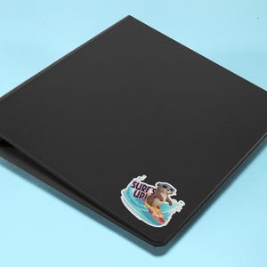 Op de afbeelding: Zwarte map met een sticker van een cartoon-otter met een zonnebril die op een surfboard surft met de tekst "Surf's Up!"