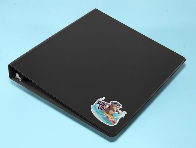 Op de afbeelding: Zwarte map met een sticker van een cartoon-otter met een zonnebril die op een surfboard surft met de tekst "Surf's Up!"