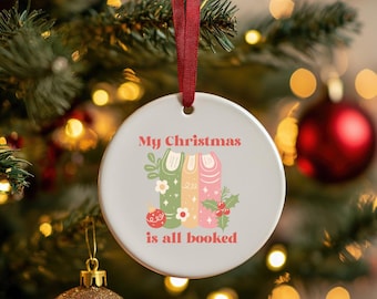 Może przedstawiać: Okrągła, biała ozdoba choinkowa z czerwoną wstążką. Ozdoba zawiera napis "My Christmas is all booked" nad grafiką trzech książek w kolorze zielonym, żółtym i różowym, z kwiatowymi akcentami.