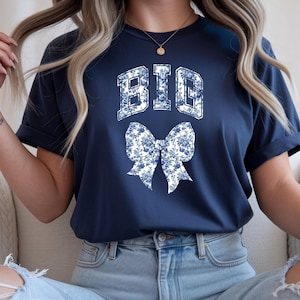 Puede incluir: Camiseta azul marino con la palabra "BIG" en blanco, con un estampado floral sobre un lazo a juego. La camiseta es de estilo cuello redondo clásico.