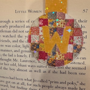 May include: A round, colorful bookmark with a large letter "W" in the center. The bookmark is made of fabric scraps sewn together in a patchwork pattern. The fabric is in shades of red, yellow, pink, and green. The bookmark is decorated with small, glittery stars.