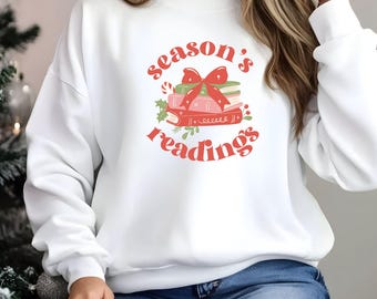 Może przedstawiać: Biała bluza z napisem "season's readings" w czerwonym skrypcie. Grafika przedstawia stos książek z czerwoną kokardą, laską cukrową i ostrokrzewem. Bluza jest z długim rękawem i okrągłym dekoltem.