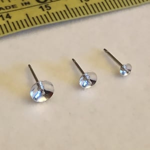 May include: Three clear, round stud earrings with silver posts are displayed on a white surface. A yellow ruler with black markings and the text "MADE IN CHINA" is in the background. The earrings vary slightly in size.
