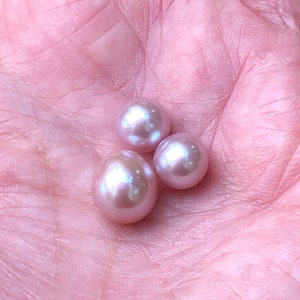 May include: Three lustrous, pink pearls of varying sizes rest in a pink palm. The pearls have a soft, iridescent sheen, reflecting light with hints of blue and green. The pearls are smooth and round, showcasing natural beauty.