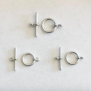 May include: Several silver-colored toggle clasps are arranged on a white surface. Each clasp consists of a straight bar with a small loop and a circular ring with a matching loop. The clasps are designed for jewelry making and crafting.