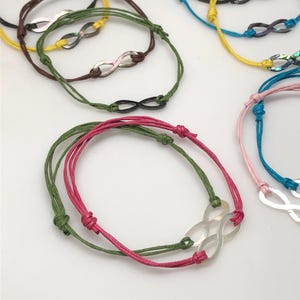 May include: Assortment of handmade infinity symbol bracelets. The bracelets feature colorful cords in shades of pink, green, yellow, and blue. Each bracelet has a metal infinity charm. The bracelets are displayed on a white surface.