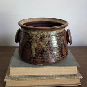 May include: A round, brown ceramic pot with two small side handles. The exterior features a mottled glaze with dark brown and tan drip patterns. The rim is wide and smooth, and the interior is a deep, solid brown color, showcasing a handcrafted finish.