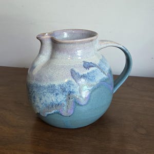 Puede incluir: Una jarra de cerámica redonda con un esmalte brillante que presenta un degradado de color desde un tono lavanda claro y blanco en la parte superior hasta una base azul cerceta profunda. Tiene un asa curva a la derecha y un pequeño pico vertedor.
