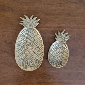 May include: Two gold pineapple-shaped serving dishes. The larger dish is on the left and the smaller dish is on the right. Both dishes have a textured pineapple design.