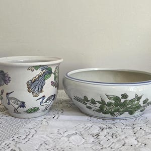 Op de afbeelding: Een witte keramieken plantenpot met een beschilderd ontwerp van kraanvogels en blauwe bloemen naast een ondiepe witte keramieken schaal versierd met een groen bloemmotief. Beide items hebben een gladde afwerking en staan naast elkaar op een ondergrond.