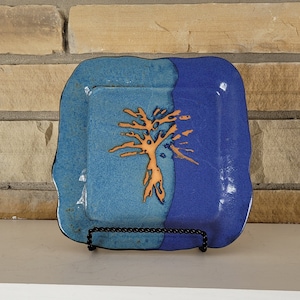 Puede incluir: Un plato cuadrado de cerámica con un diseño de árbol. El plato está dividido en dos secciones, una azul claro y otra azul oscuro. Un diseño de árbol naranja está en el centro. El plato está sobre un soporte de metal negro.