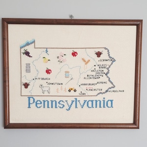 May include: Framed cross-stitch map of Pennsylvania, featuring embroidered icons representing local products and landmarks. The map is in a dark brown wooden frame, with the state name stitched in blue.