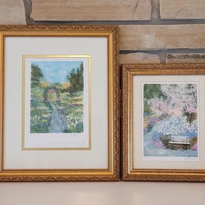 Könnte beinhalten: Zwei gerahmte Landschaftskunst-Drucke. Der größere Druck zeigt einen Gartenweg mit Blumen und Bäumen, goldgerahmt. Der kleinere Druck zeigt eine Bank unter einem blühenden Baum, ebenfalls in einem goldenen Rahmen. Beide Drucke haben ein weißes Passepartout.