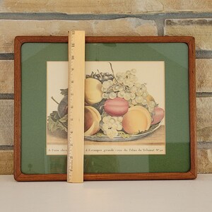 May include: A framed print of a still life featuring a bowl of fruit. The bowl contains peaches, plums, and grapes. The print is in a wooden frame with a green mat. The text below the image reads "A l'Etampe grande cour du Palais du Trianon N. 30."