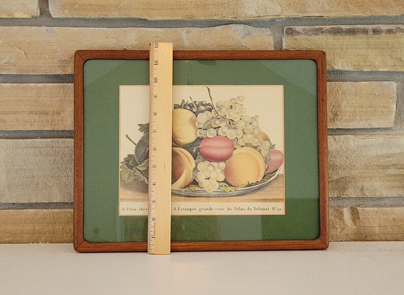 May include: A framed print of a still life featuring a bowl of fruit. The bowl contains peaches, plums, and grapes. The print is in a wooden frame with a green mat. The text below the image reads "A l'Etampe grande cour du Palais du Trianon N. 30."