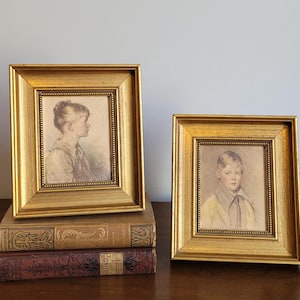 May include: Two gold framed portraits of children. The portrait on the left is of a girl with light brown hair, wearing a white dress. The portrait on the right is of a boy with light brown hair, wearing a white shirt and a dark tie.