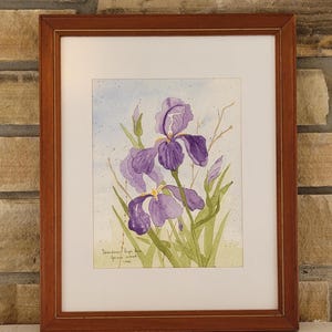 Peut inclure: Peinture à l'aquarelle d'iris violets avec des feuilles et des tiges vertes, sur un fond bleu clair et jaune. L'œuvre est encadrée dans un cadre en bois brun foncé et signée par l'artiste.