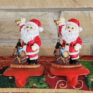 Puede incluir: Un par de sujetalibros de Santa Claus rojos y blancos con una campana en cada mano. Las figuras de Santa Claus sostienen una bolsa de regalos, un osito de peluche y un árbol de Navidad. Los sujetalibros están sobre una superficie con estampado rojo y verde.