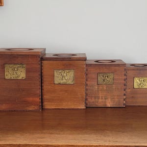 May include: Four wooden boxes of varying sizes, arranged in a row. Each box is a warm brown color and features a brass-colored rectangular plaque with an embossed design. The boxes have a square shape with a circular opening on top.